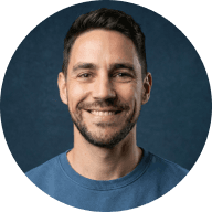 Smiling man with short dark hair and beard wearing a gray shirt against a dark blue background.